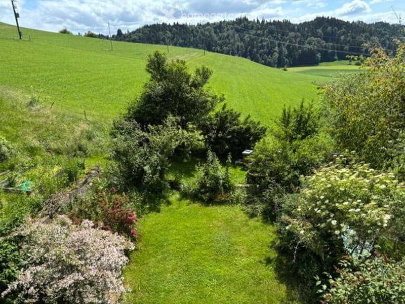 A lush, green garden with well-maintained lawn, various shrubs, and trees, surrounded by rolling hills and a forested area in the background.