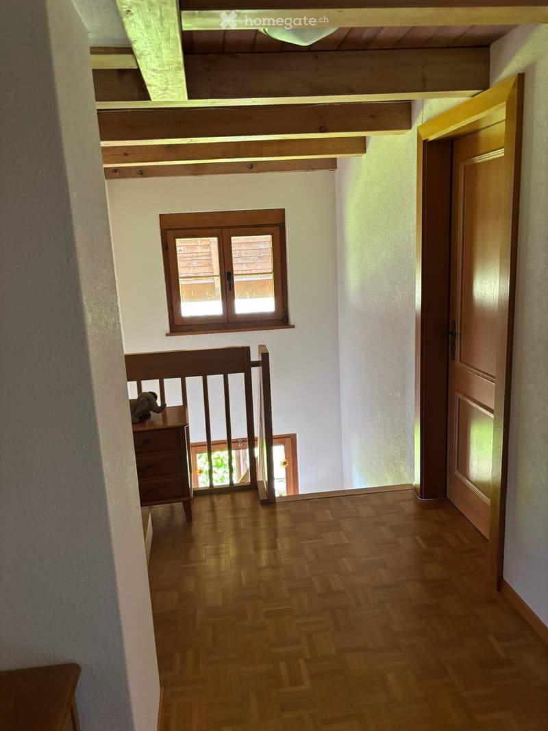 Wooden hallway with herringbone flooring, white walls, and a small window. Features a wooden railing leading to an upper level and a closed door on the right.
