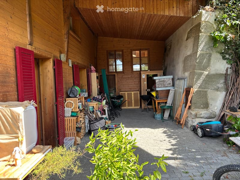 Wooden outdoor area with storage items, tools, and a covered refrigerator, surrounded by greenery and stone walls.