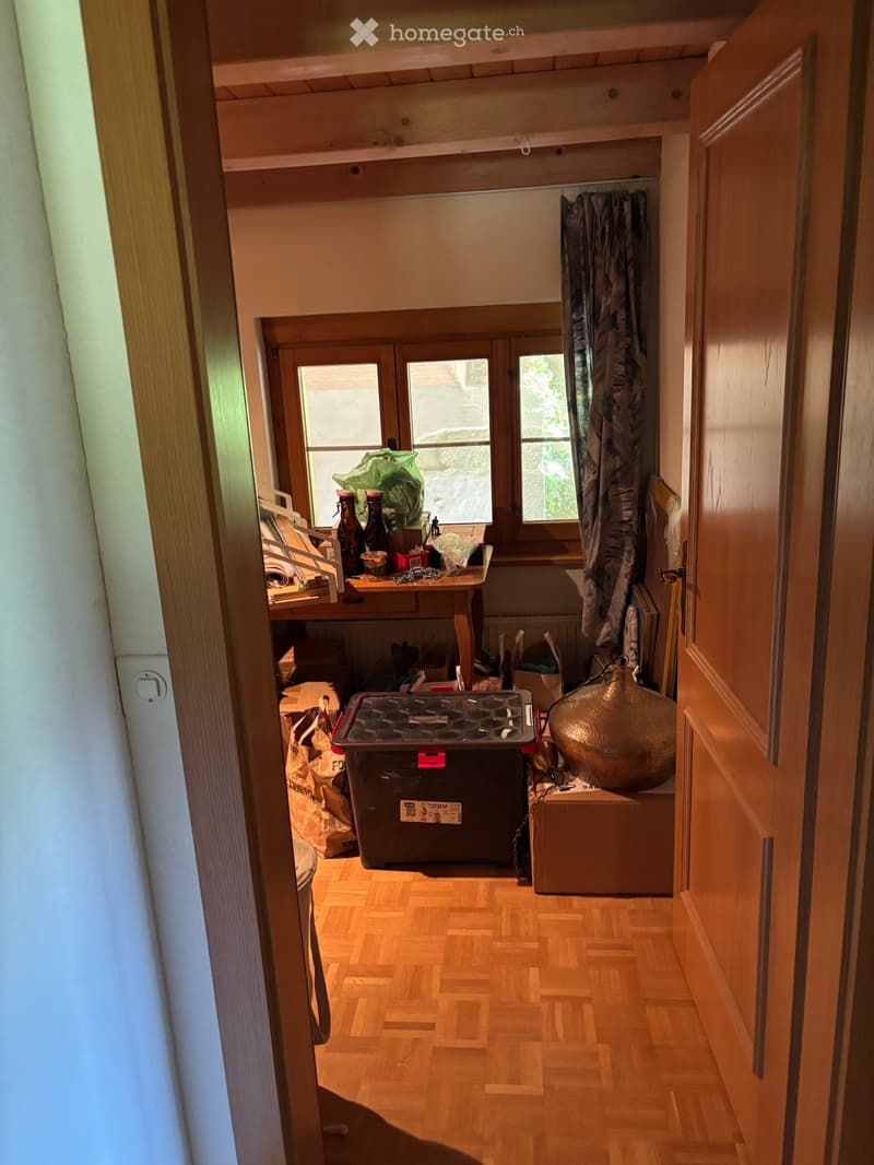 A small, cluttered room with wooden flooring, a table with various items, a curtain, and storage boxes.