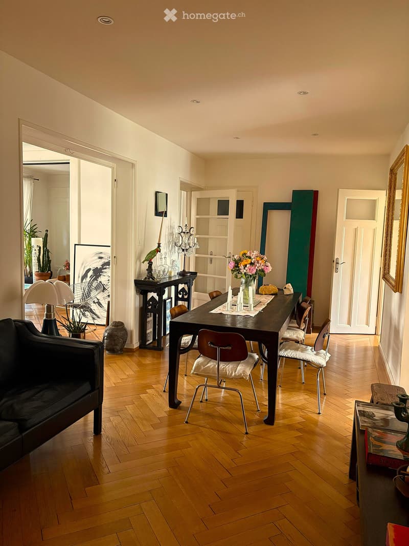 Bright, open living and dining area with wooden flooring, a black sofa, a dining table with chairs, a lamp, and decorative items including a vase with flowers and a mirror.