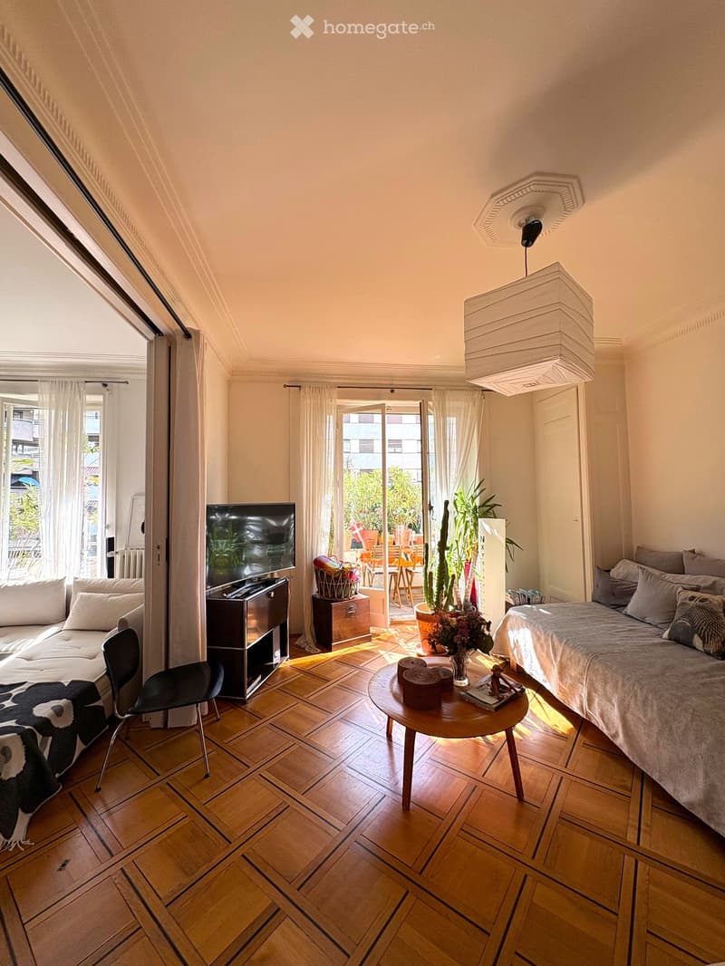 A well-lit room with wooden flooring, a sofa, a bed, a television, a round table, and a chair. The room features large windows with sheer curtains and a view of a garden area.