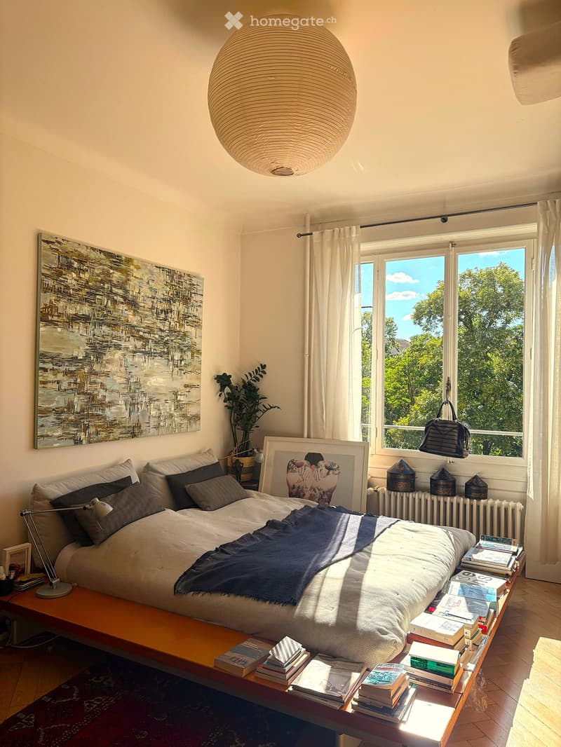 A modern bedroom with a large bed, pillows, a blanket, a bedside table with books and a lamp, a painting on the wall, a plant, and a large window with curtains.