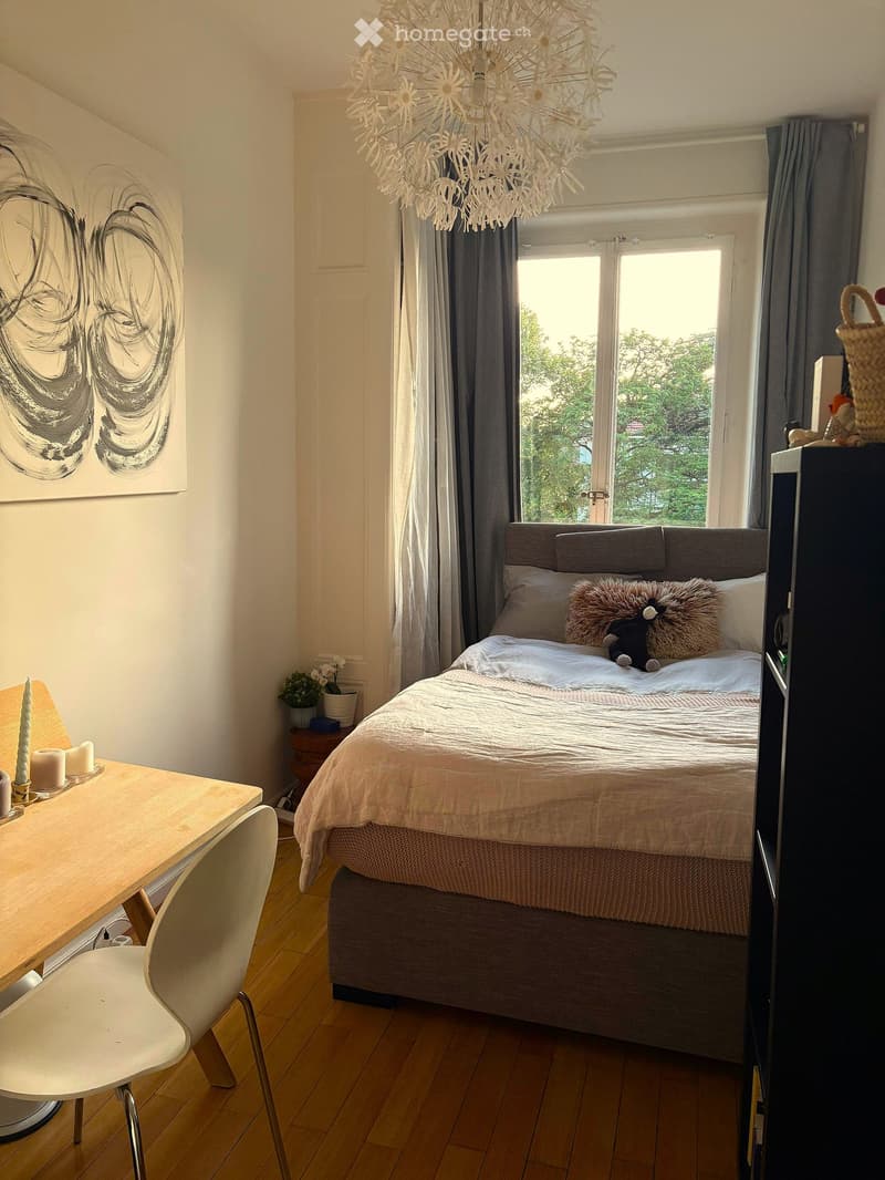 Cozy bedroom with a large bed, wooden floor, white curtains, and a modern chandelier. Includes a small table with candles, a chair, and a potted plant on a side table.