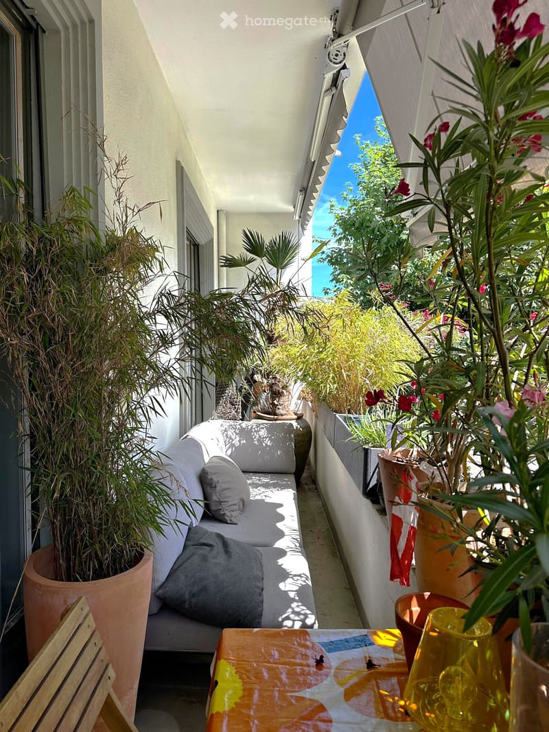 Balcony with a grey sofa, wooden table, and various potted plants, offering a scenic view of greenery and the sea.
