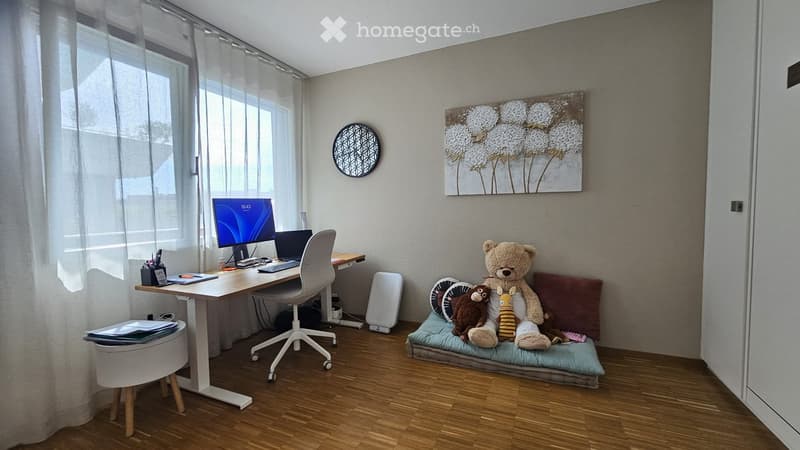 A modern, well-lit room with a wooden floor, a desk with a laptop and monitor, a comfortable sofa with pillows and stuffed animals, and a large window with sheer curtains.