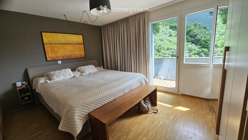 Modern bedroom with a large bed, nightstand, bench, and a painting on the wall. The room features wooden flooring and large windows with a view of greenery.
