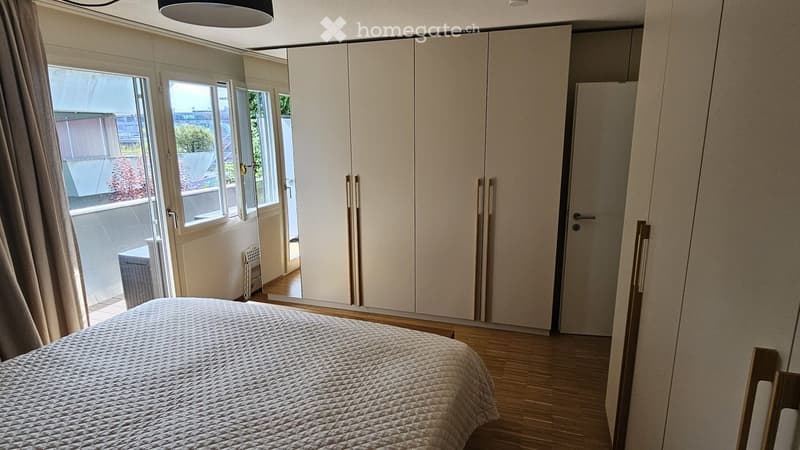Modern bedroom with a neatly made bed, large built-in wardrobe, and a large window with a curtain leading to a balcony.