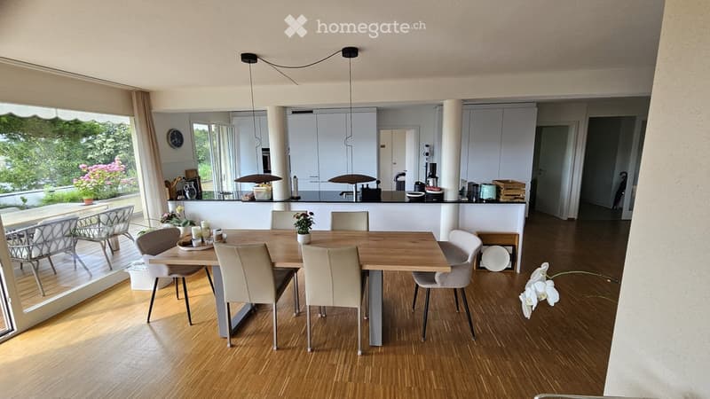 Bright dining area with a wooden dining table and six chairs, modern kitchen with white cabinets and various appliances, pendant lights, and a view of a garden through large windows.