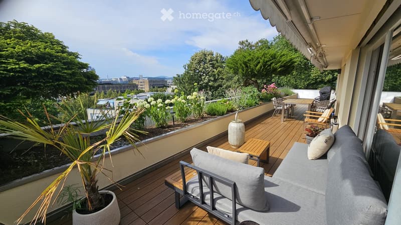 A spacious, well-maintained balcony with wooden decking, outdoor furniture including a sofa, small tables, and chairs, surrounded by lush greenery and potted plants. The view includes a cityscape with trees and buildings.