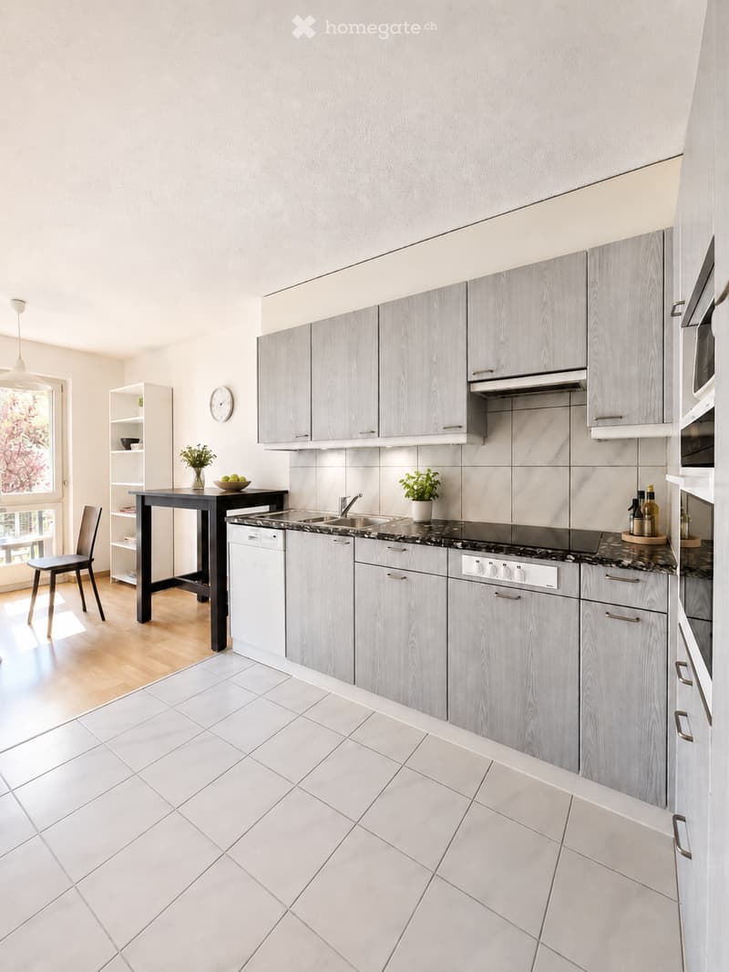 Modern kitchen with grey cabinets, white tiled floor, marble countertop, sink, oven, dishwasher, table, chair, and decorative items.