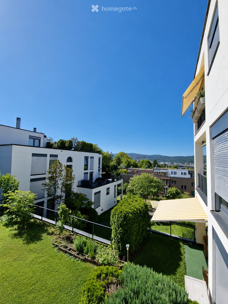 A well-maintained garden area with green grass, shrubs, trees, and a small pathway. The garden is enclosed by a metal fence and features a small seating area with a table and chairs under a striped awning. The background shows modern apartment buildings a
