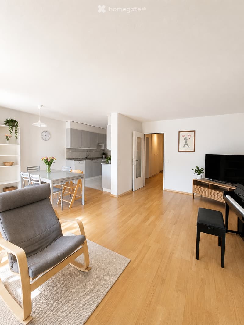 Bright, open-plan living and dining area with light wooden flooring, a grey sofa, a dining table with chairs, a piano, a TV, and a small kitchen area with cabinets and appliances.
