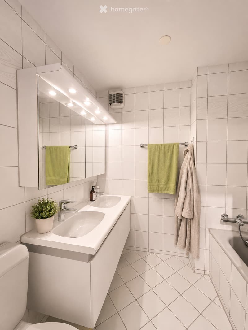Modern bathroom with white tiled walls and floor, double sink vanity, large mirror with built-in lighting, towels, and a small potted plant on the counter.
