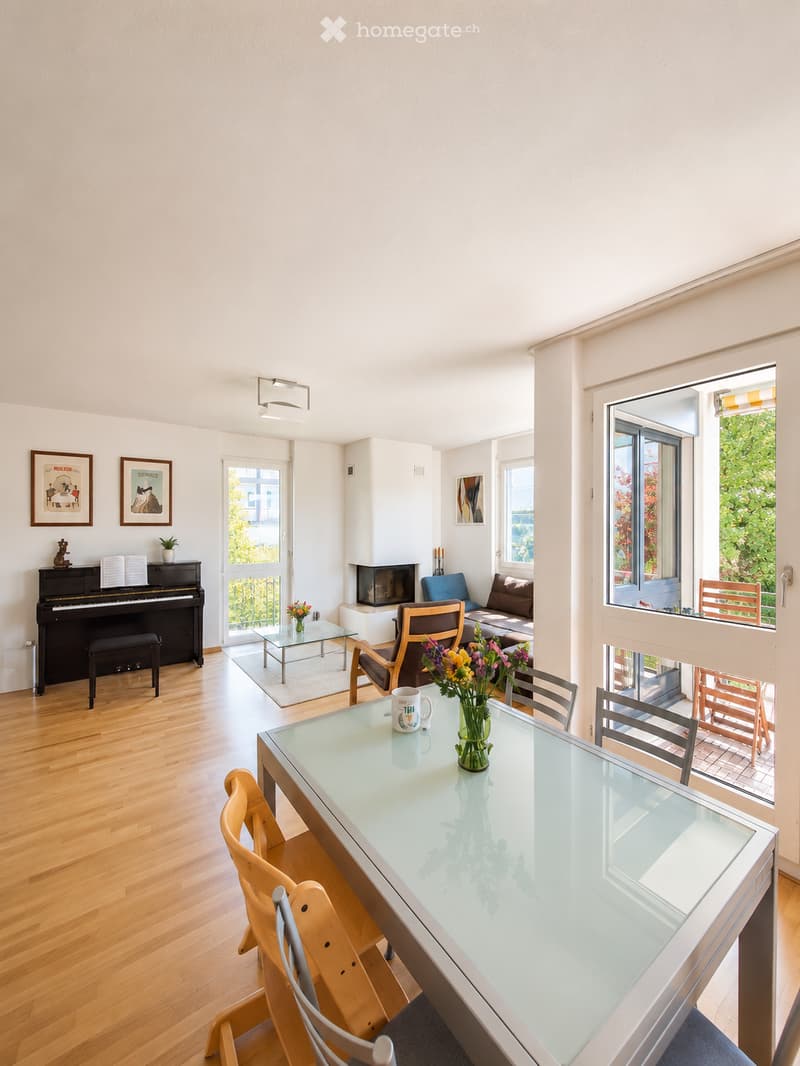 Bright, open living and dining area with wooden flooring, large windows, and a balcony. Features a piano, sofa, armchair, fireplace, glass coffee and dining tables, and chairs.