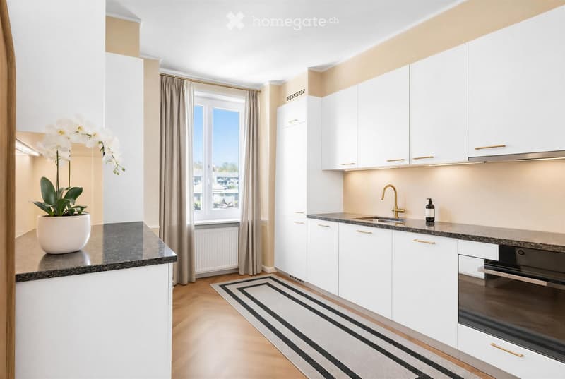 Modern kitchen with white cabinets, granite countertops, a sink with a gold faucet, and a built-in oven. A potted orchid sits on the counter, and a striped rug is on the floor.
