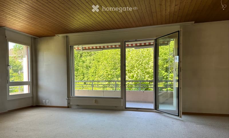 Empty room with wooden ceiling, large windows, and sliding glass doors opening to a balcony.