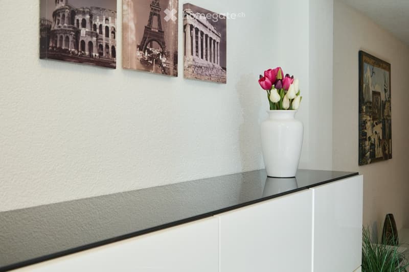 A hallway with a glass console table, a white vase with flowers, and several framed pictures on the walls.
