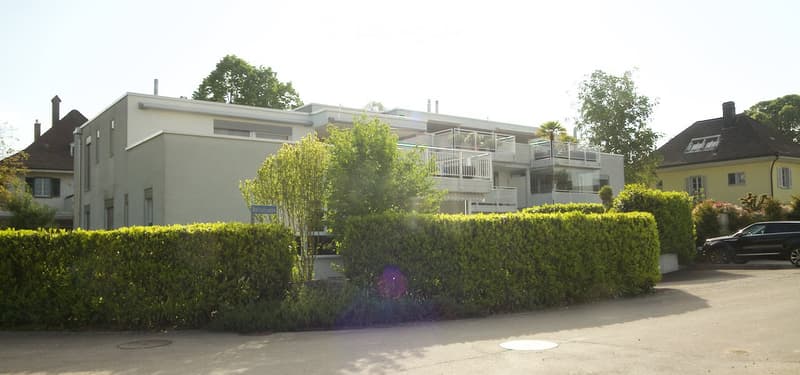 Modern two-story building with a balcony, surrounded by greenery and a hedge in front.
