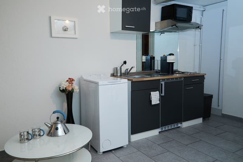 Compact kitchen with dark cabinets, stainless steel appliances, a small table with teapot and cups, and a vase with flowers.
