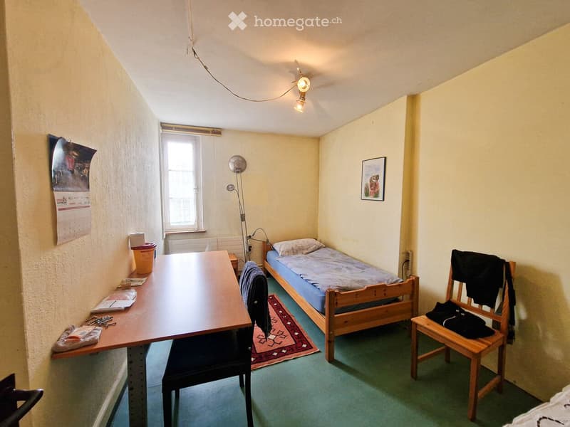 Small furnished room with a single bed, desk, chair, and a small table. Features a slanted ceiling and a window letting in natural light.