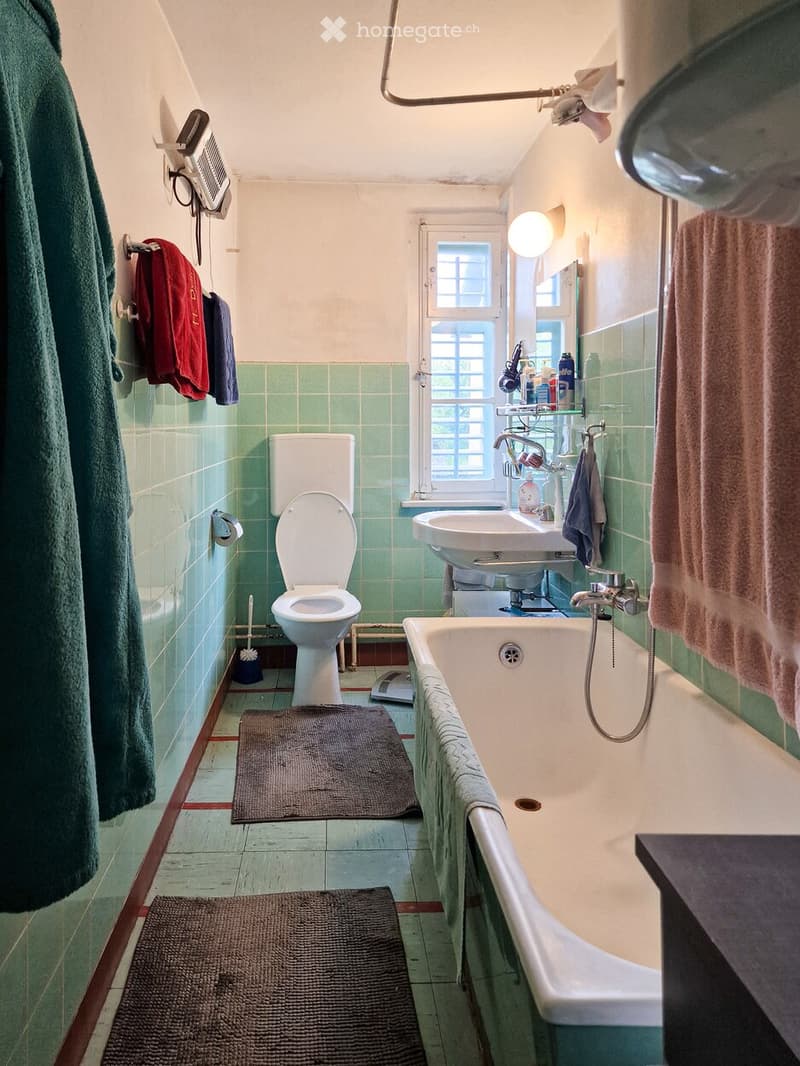 Bathroom with green tiled walls, a toilet, a bathtub, a sink, a mirror, towels, a bathrobe, a brush, and a scale.