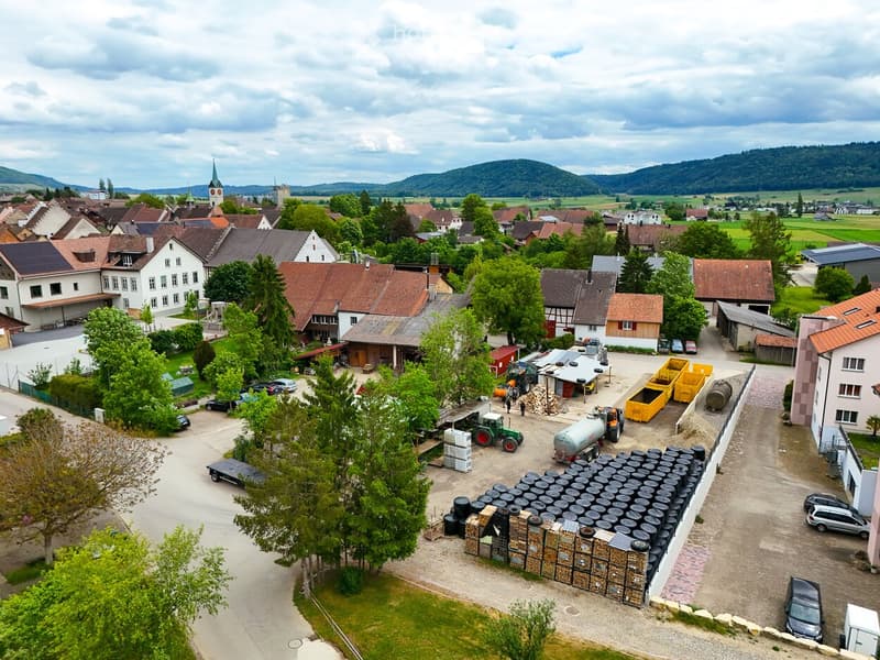 Aerial view of a rural village with scattered houses, a church tower, and a farm area with vehicles, storage containers, and agricultural equipment.