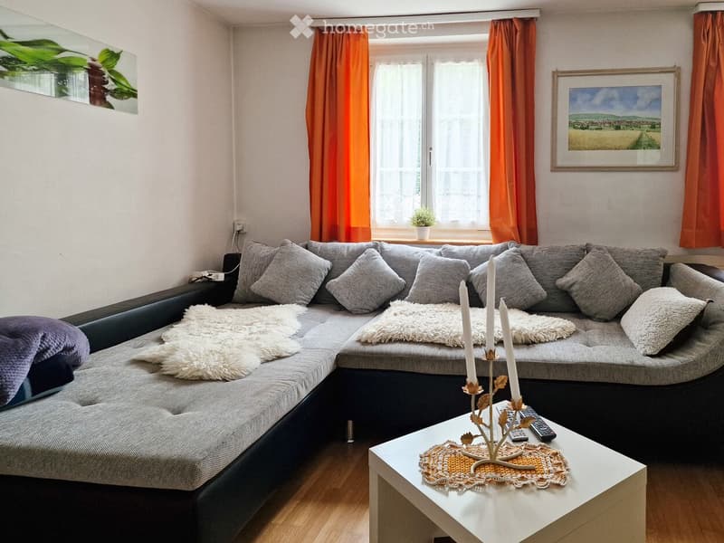 Modern living room with a grey sectional sofa, white coffee table, candle holder, and decorative pillows. Large windows with orange curtains and a small plant on the windowsill.