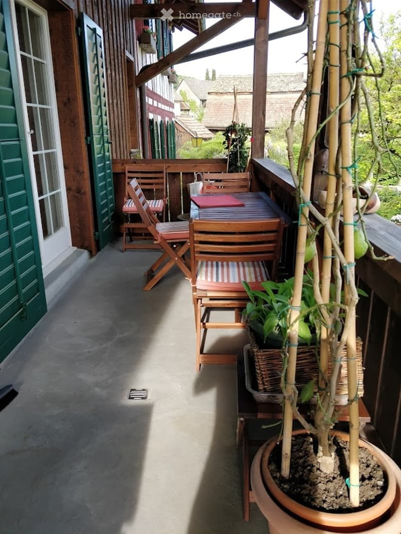 Wooden balcony with outdoor furniture including chairs and a table, surrounded by potted plants and greenery.