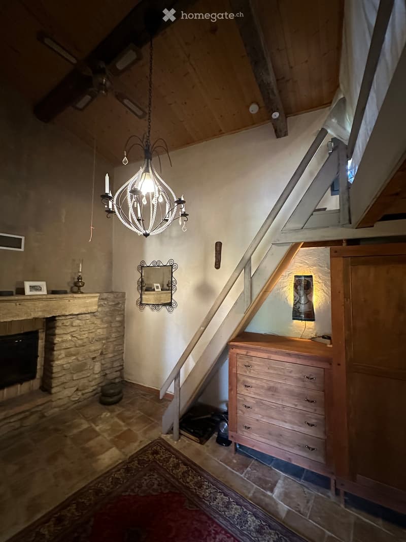 Cozy attic room with exposed wooden beams, a stone fireplace, a chandelier, a wooden drawer unit, and a patterned rug.