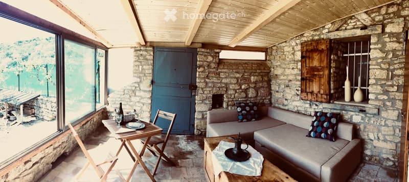 Cozy, rustic living space with stone walls, wooden ceiling beams, and large windows. Features a sofa with patterned cushions, a small wooden table with chairs, and decorative vases on a stone shelf.