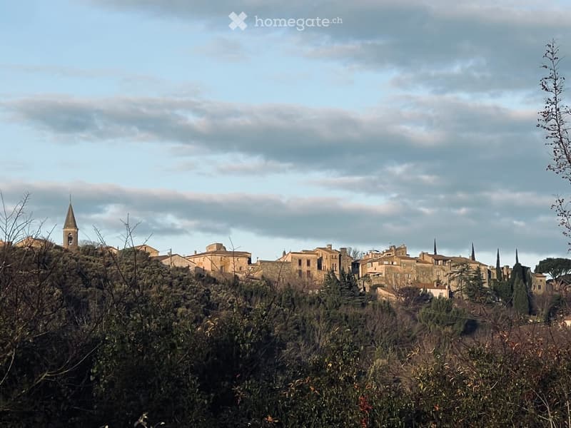 A picturesque village with stone houses, a church tower, and surrounding greenery under a partly cloudy sky.