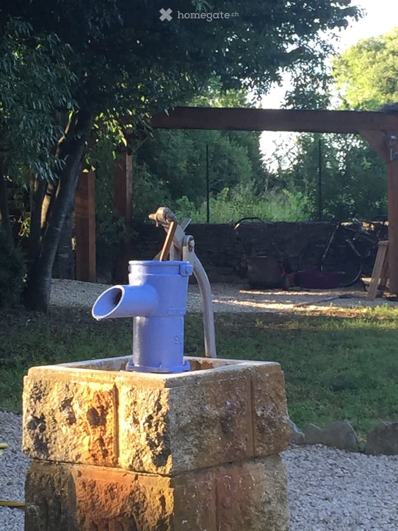A stone water fountain with a blue spout and handle, situated in a garden area with gravel ground and trees in the background.