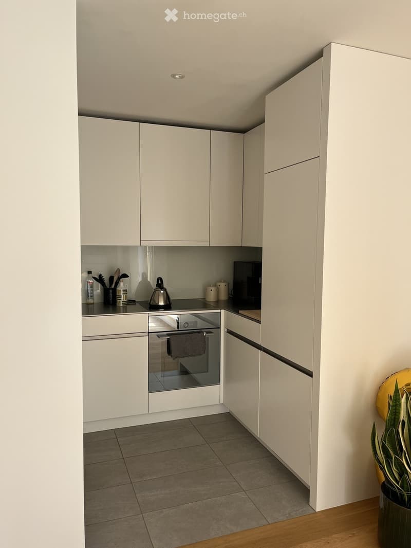 Modern kitchen with white cabinets, stainless steel oven, countertop items including a teapot, coffee maker, and various bottles.