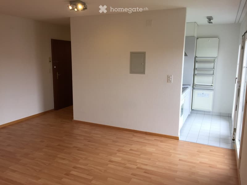 Empty room with wooden flooring, white tiled kitchen area, and a refrigerator. No furniture or personal items present.