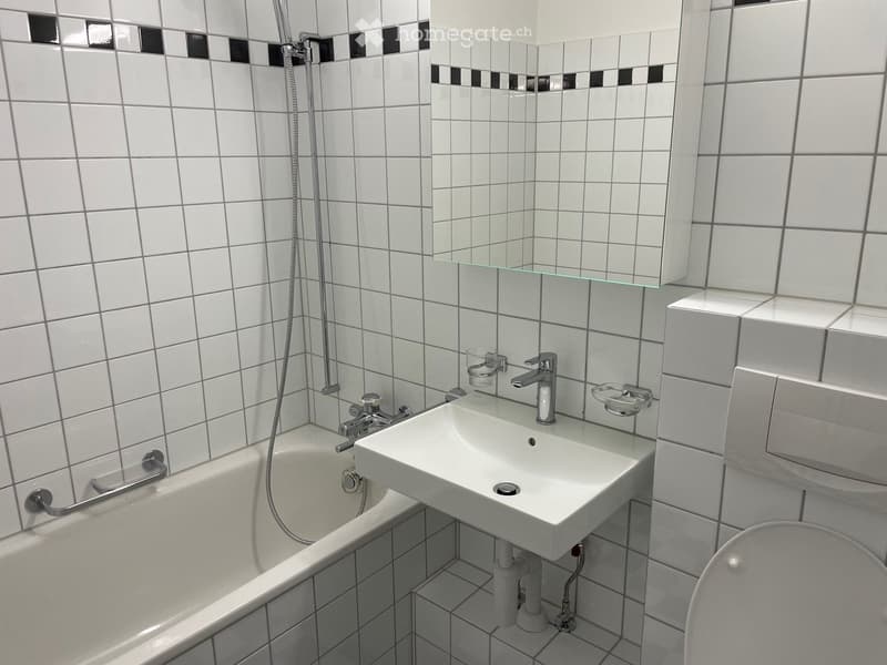 Modern bathroom with white tiled walls, a bathtub, a sink with a modern faucet, a mirror, and a toilet. The tiles feature a black and white pattern at the top.