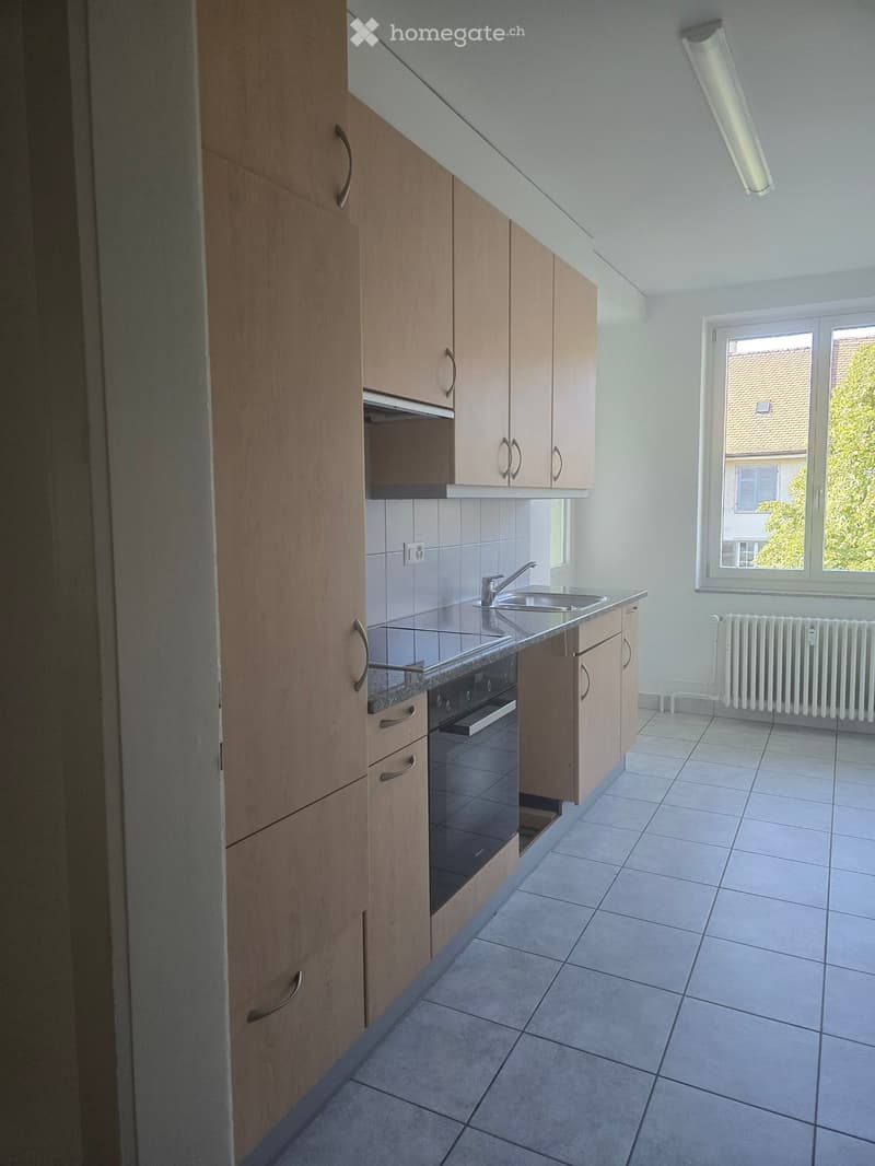 white tiled floor, wooden cabinets, stainless steel countertop, double sink, black oven and stove, white radiator, window with view outside