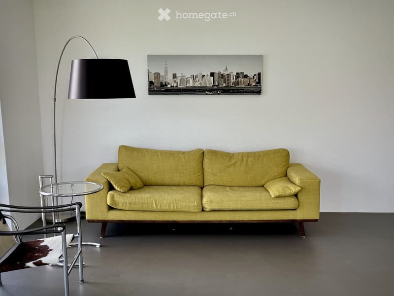Modern living room with a yellow sofa, glass coffee table, sleek chair, floor lamp, and a cityscape picture on the wall.