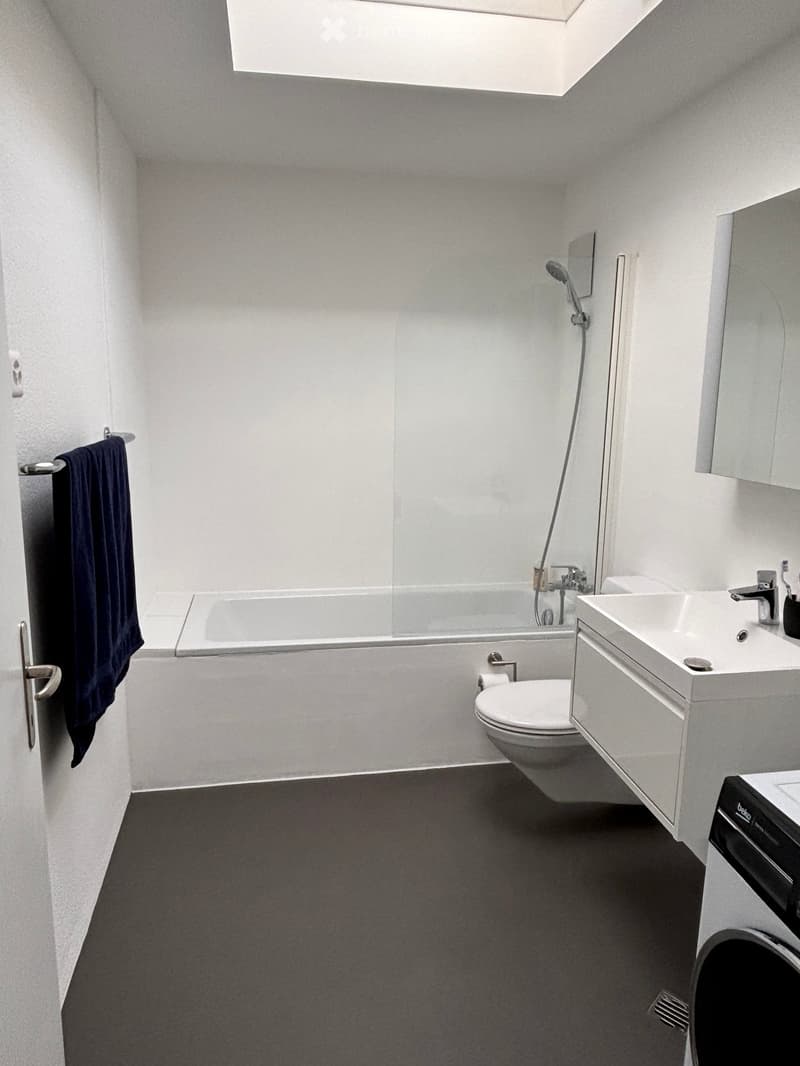 Modern bathroom with white walls, a bathtub, toilet, sink, mirror, and a towel hanging on a rack.