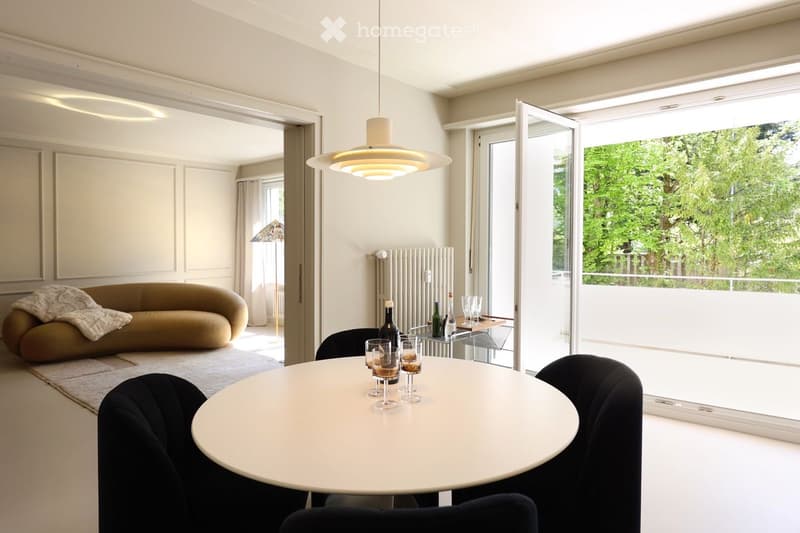 Dining room with round table, black chairs, white walls, large windows, balcony, couch, wine glasses, bottle, radiator