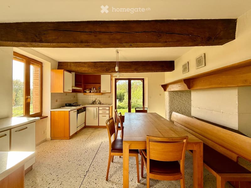 Bright kitchen with wooden beams, white cabinets, a central dining table with chairs, and a large window overlooking greenery.
