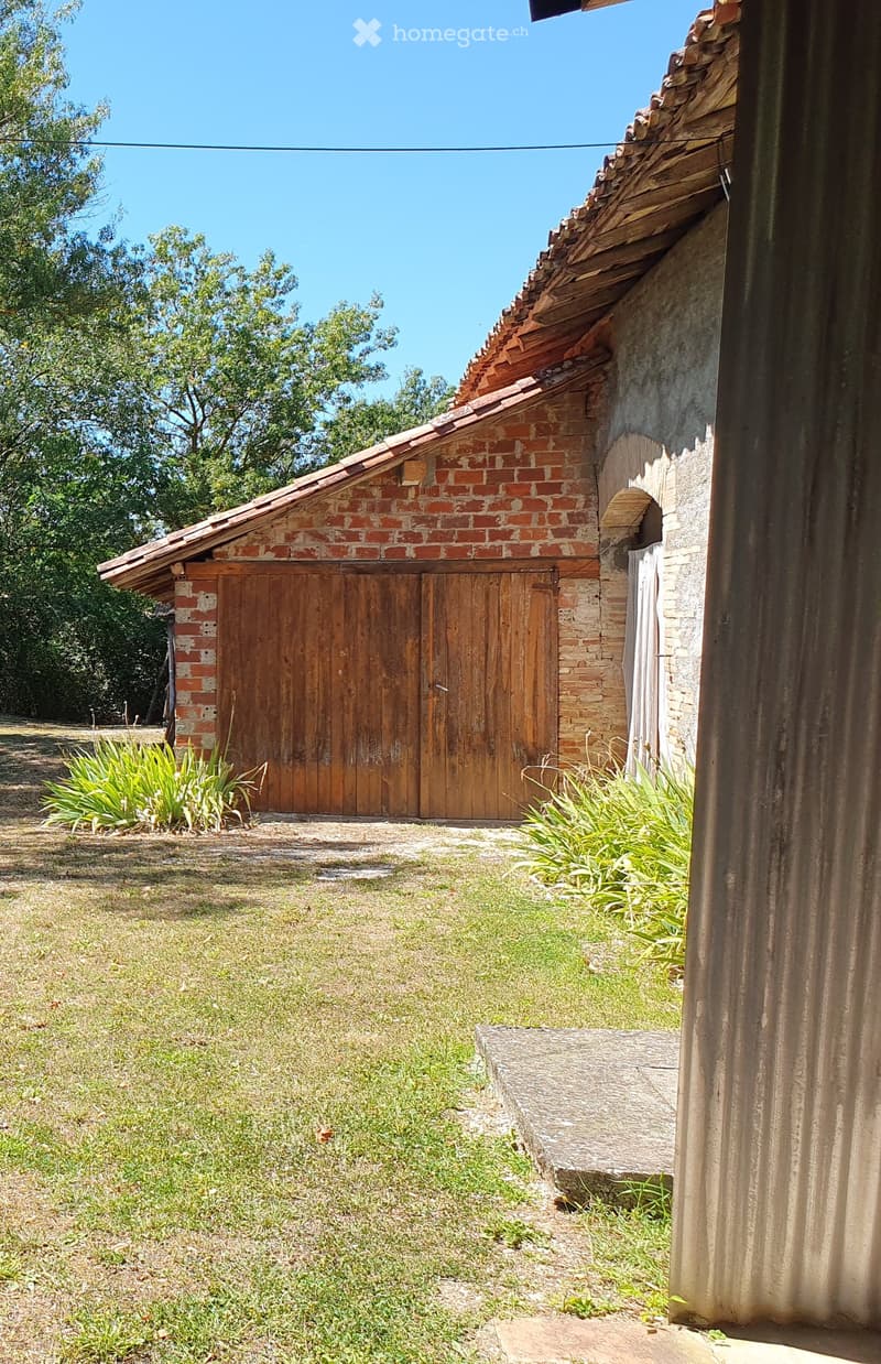 A small brick outbuilding with a wooden door, surrounded by grass and plants, with a stone step leading up to it.