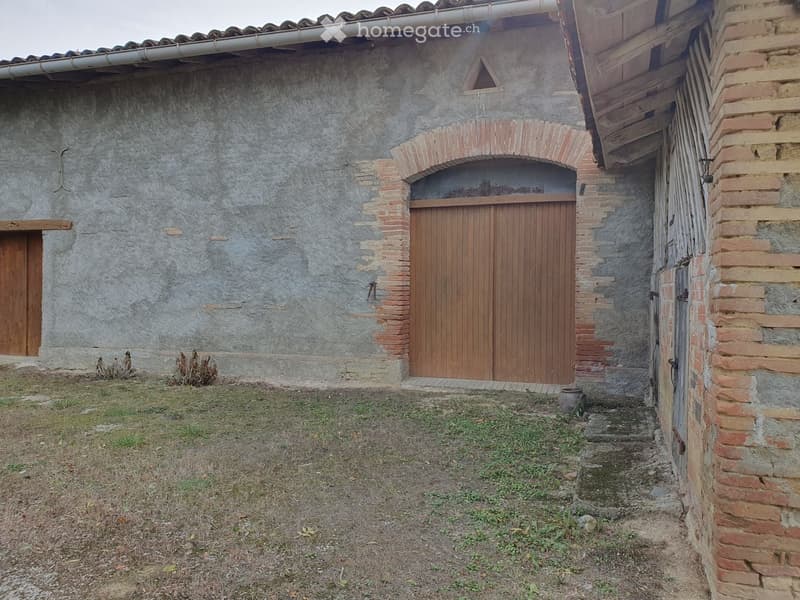Exterior view of a rustic stone building with wooden doors and a small grassy area in front.