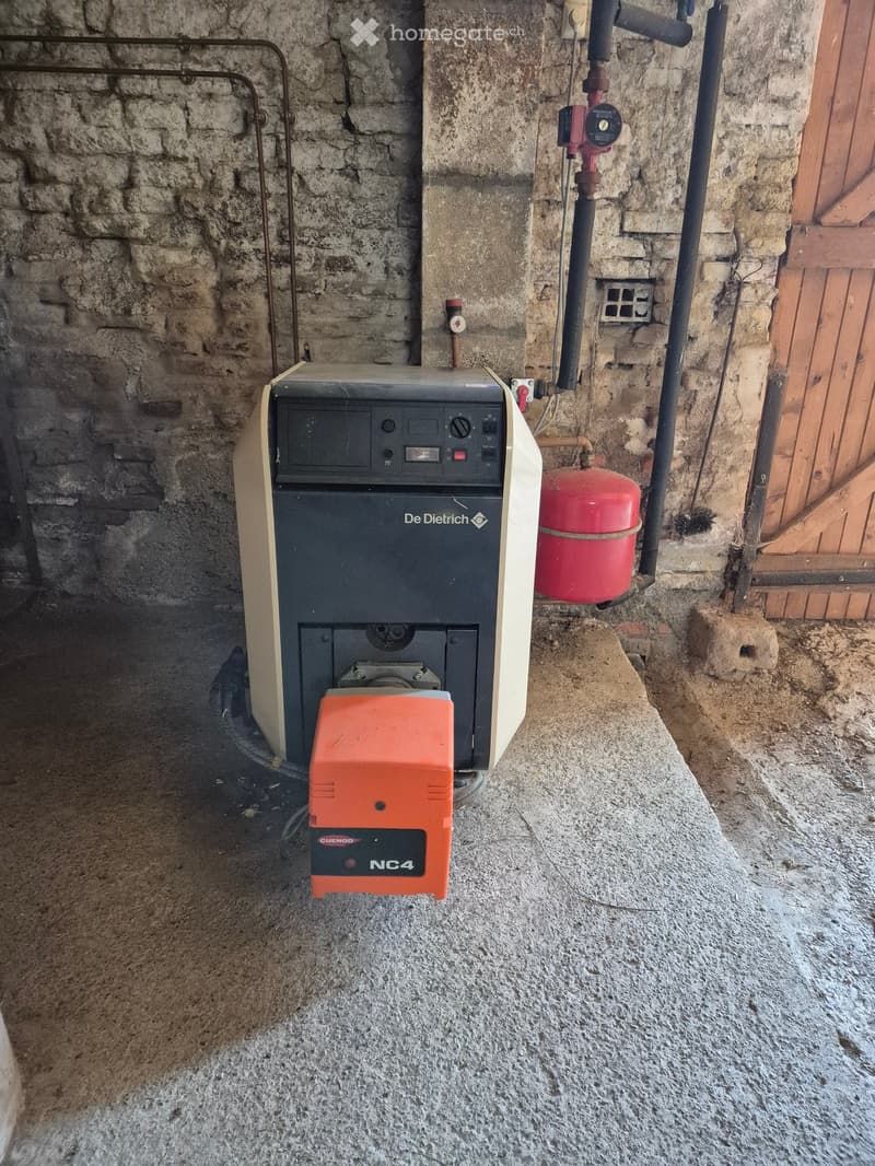 A water heating system with a De Dietrich water heater, a red gas cylinder, and an orange control unit labeled "NC4" mounted on a stone wall in a utility area.