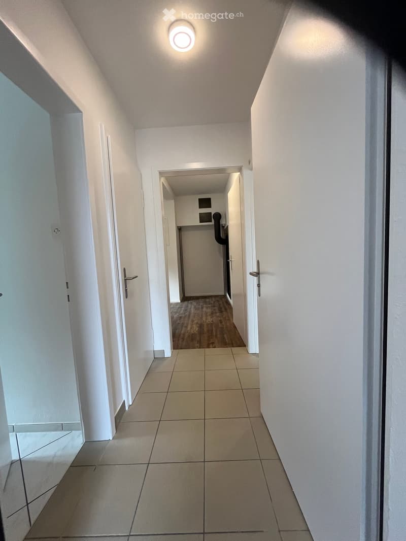 Clean, modern hallway with tiled flooring, white walls, and several doors leading to other rooms. A round ceiling light illuminates the space.