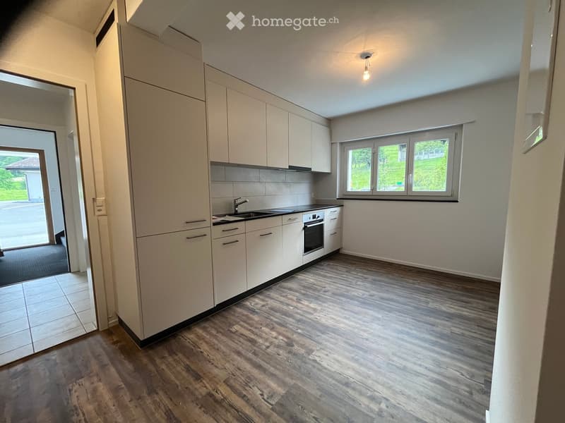 Modern kitchen with light-colored cabinets, a sink, and an oven. The floor is made of wood, and there is a large window with a view of greenery outside.