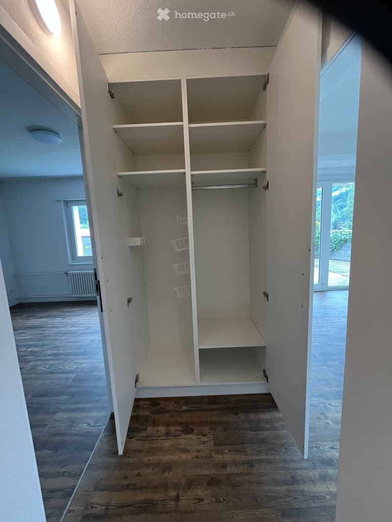 Empty room with wooden flooring, white walls, built-in closet with empty shelves, and a window letting in natural light.