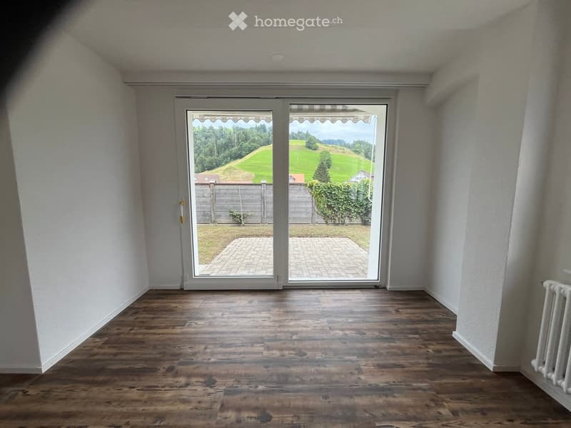 Empty room with wooden flooring, white walls, and large sliding glass doors leading to a patio and green landscape.