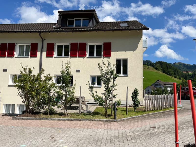 Two-story house with beige walls, red shutters, and multiple windows. Small garden area with trees and a bench in front.