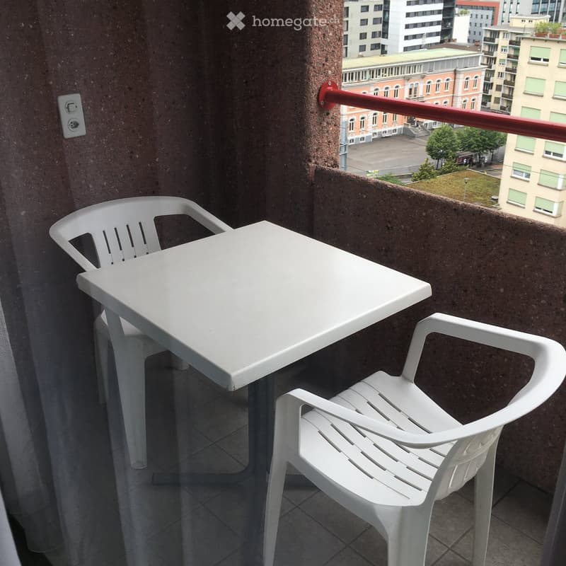 Balcony with a white plastic table and chair, overlooking a cityscape with buildings and greenery.
